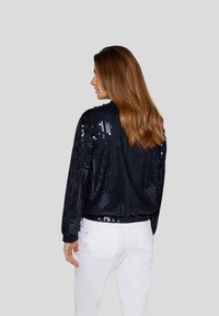 Navy-Bomberjacke mit Pailletten, ausgestattet mit einem gerippten Kragen und Bündchen, im taillierten Design. Das Model trägt weiße Jeans und präsentiert die Rückansicht.