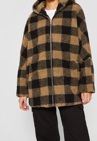 Fleece hættejakke i et brunt og sort ternet mønster, med en frontlynlås og løs pasform, udstyret med sænkede skuldre og struktureret stof.