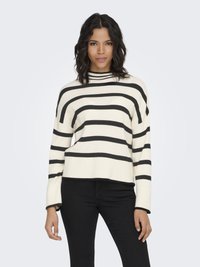 Woman with wavy dark hair wearing a cream sweater with wide black stripes and black pants, standing against a plain light background.