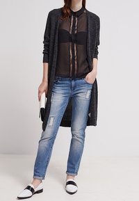 Lang grå cardigan over en sort gennemsigtig skjorte med knapper, slidte lyseblå jeans og sorte og hvide loafers. Clutch i hånden.