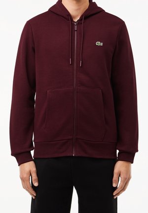 Mann trägt einen dunklen bordeauxroten Kapuzenpullover mit Reißverschluss, vorderen Taschen und einem kleinen grünen Krokodillogo auf der Brust.