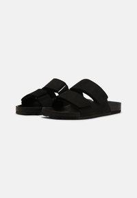 Sandales noires avec des doubles straps réglables, semelles plates et texture lisse. Fabriquées en matériau semblable à du daim avec une semelle intérieure large.