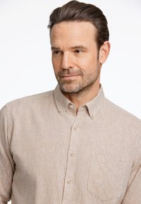 Camisa beige de botones con una tela texturizada, que presenta un bolsillo y un cuello clásico. Los botones son ligeros y redondos. Diseño simple y funcional.