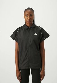 Casaco atlético preto com fecho à frente, mangas curtas em forma de copo, com detalhes brancos e o logótipo da Adidas na área do peito esquerdo.