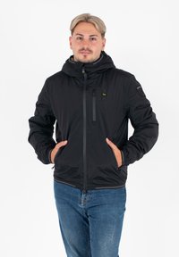 Giacca nera con zip e cappuccio, dotata di polsini elastici, due tasche laterali e una piccola tasca sul petto con dettaglio del logo. Texture liscia.