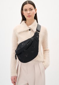 Fekete szövet hordszíj, virágos gyöngyökkel díszítve, széles fekete pánttal rögzítve, bézs kötött pulóver fölött viselve.