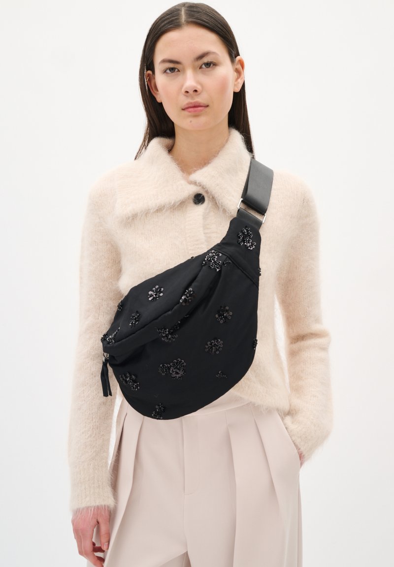 Fekete szövet hordszíj, virágos gyöngyökkel díszítve, széles fekete pánttal rögzítve, bézs kötött pulóver fölött viselve.
