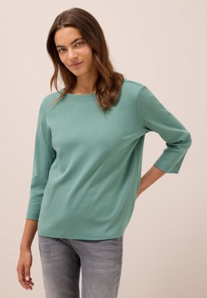 Femme aux longs cheveux bruns portant un haut en jersey teal à manches trois-quarts et un jean gris délavé, posant avec une main derrière le dos.