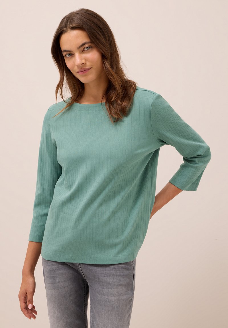 Femme aux longs cheveux bruns portant un haut en jersey teal à manches trois-quarts et un jean gris délavé, posant avec une main derrière le dos.