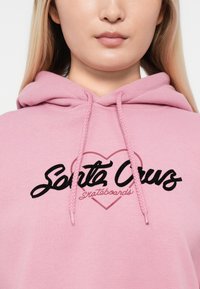 Felpa rosa con cappuccio e lacci, con la scritta "Santa Cruz" in carattere nero e "skateboards" in testo più piccolo sotto, su un tessuto morbido.