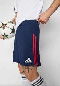 Marineblauwe sportshorts met rode en witte gestreepte accenten, met een elastische tailleband en het Adidas-logo in het wit aan de zijkant.