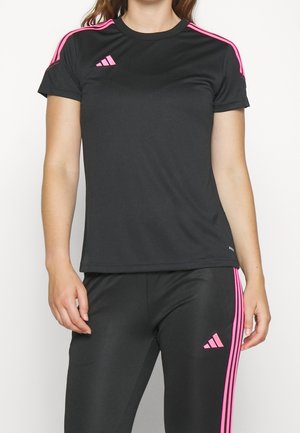 Femme portant une tenue de sport noire avec logo Adidas rose et des bandes sur les manches et le pantalon, se tenant devant un fond uni.