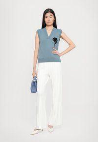 Pull sans manches en tricot bleu avec un décolleté en V et un détail floral noir, assorti à un pantalon large plissé blanc et des escarpins beiges à bout pointu.