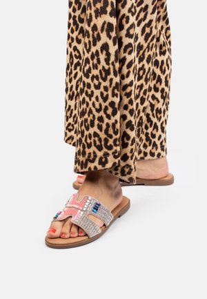 Beaded slide sandals with a cross design, featuring pink and blue accents, set against a light tan sole. Leopard print fabric drapes nearby.