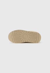 Suela de zapato beige con patrones de tracción geométricos y un pequeño logotipo circular cerca del dedo del pie sobre un fondo blanco liso.