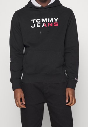 Personne portant un sweat à capuche noir avec poche avant et logo "TOMMY JEANS" en lettres blanches et rouges sur la poitrine.