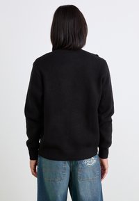 Schwarzer gestrickter Pullover mit Rundhalsausschnitt, langen Ärmeln und gerippten Bündchen, kombiniert mit lockeren blauen Jeans aus Denim mit einem Logopatch.