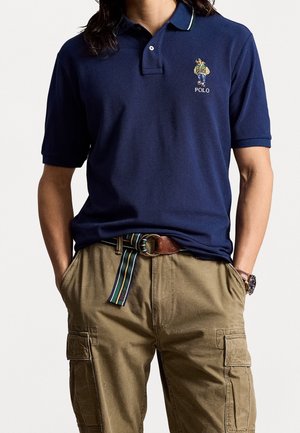 Mann trägt ein marineblaues Poloshirt mit gesticktem Bärenlogo, beige Cargo-Hose, gestreiften Gürtel und Armbanduhr, die Hände in den Taschen.