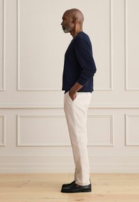 Un homme est de profil, vêtu d'un pull bleu marine, d'un pantalon beige clair et de mocassins noirs, les mains dans les poches, devant un mur beige à panneaux.
