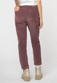 Person, der dunkelviolette Cordhosen mit hochgekrempeltem Saum und beigen Buffalo-Plateauschuhen trägt, steht vor einem schlichten hellgrauen Hintergrund.