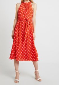 Röd klänning med prickar, halterneck-design, åtdragen midja och flödande kjol. Tillverkad av lätt tyg med en mjuk struktur.