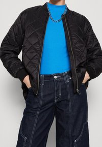 Schwarze, gesteppten Bomberjacke mit gerippten Bündchen, Reißverschluss und glatter Textur. Kombiniert mit einem hellblauen, gerippten Oberteil und dunklen Jeanshosen.