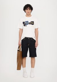T-shirt blanc avec des motifs colorés brodés sur la poitrine, shorts en jean foncé, baskets blanches et un sac fourre-tout marron avec une finition brillante.