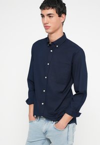 Laivastonsininen, napitettava pitkähiha-paita, jossa on rintatasku ja valkoiset napit. Kangas näyttää sileältä ja tasalaatuiselta.