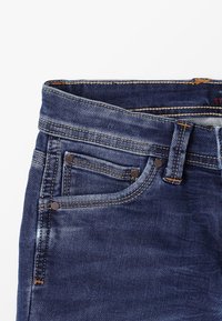 Mörkblå denimjeanssektion som visar framfickan, myntfickan, bältesöglan och detaljerad sömnad mot en vit bakgrund.