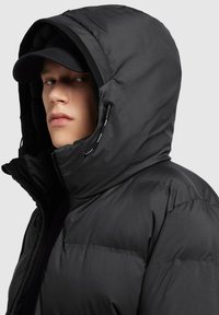 Sort pufferjakke med en stor hætte, struktureret stof og justerbare snore; båret med en sort kasket, tæt på, sideprofil.