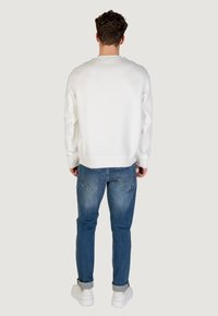 Hombre de pie de espaldas, vestido con un sudadera blanca, jeans azules con los puños enrollados y zapatillas blancas, sobre un fondo liso.