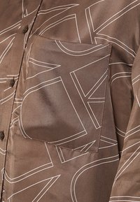 Ruskea paita, jossa on valkoisia abstrakteja viivakuosisia kuvioita, rintatasku ja ruskeat napit sileässä kankaassa.