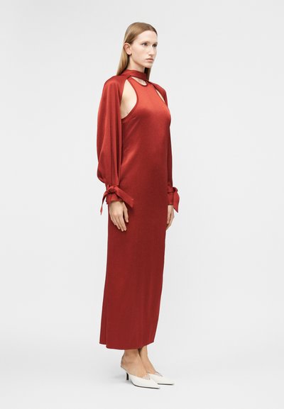 Γυναίκα που φοράει ένα αμάνικο κόκκινο σατέν φόρεμα με ροές δεμένες στα μανίκια και λευκά μυτερά παπούτσια στιλέτο, στέκεται μπροστά σε ένα απλό φωτεινό φόντο.