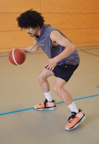 Basketballspeler die een bal dribbelt, gekleed in een grijs mouwloos shirt, zwarte shorts en oranje sneakers met zwart-witte accenten.