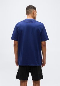 Άνδρας με κοντά μαλλιά, φορώντας φαρδιά μπλούζα ναυτικού μπλε με κοντά μανίκια και μαύρα σορτς, στέκε ται γυρισμένος με την πλάτη προς το μέρος μας σε μια απλή φόντο.