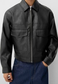 Zwarte jas van nep leer met een ritssluiting aan de voorkant, een puntige kraag, twee borstzakken en een cropped ontwerp. Draagt over een blauwe spijkerbroek.