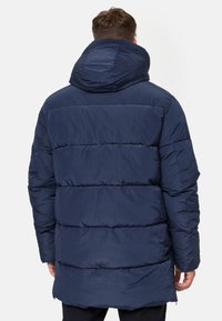 Veste rembourrée bleu marine avec un col haut et une capuche, présentant un design matelassé et des coutures contrastées. Texture lisse, isolée pour la chaleur.