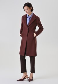 Cappotto in lana bordeaux con rever a punta, chiusura a bottoni e tasche laterali, abbinato a una blusa azzurra chiara e pantaloni neri a fantasia.