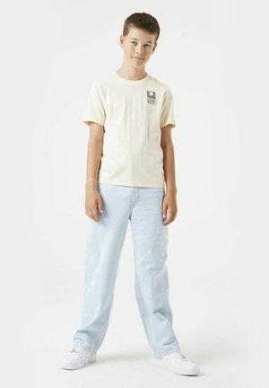 Camiseta de algodón amarillo claro con un gráfico en el pecho, combinada con jeans de mezclilla azul claro y zapatillas blancas, de pie contra un fondo blanco.