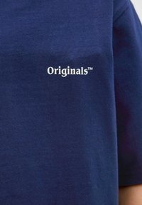 Tumed sinine puuvillane T-särk, mille vasakus rinnaku osas on valge "Originals™" logo, sile tekstuur ja klassikaline lühikeste varrukate disain.