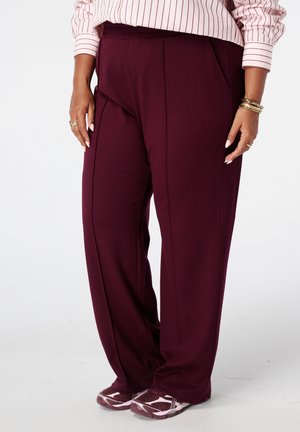 Femme portant un pantalon large violet foncé avec des coutures à l'avant, des baskets roses et blanches, une chemise rose clair à rayures, des bracelets et des bagues.