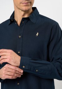 Tums zilgana garām piedurknēm ar pogu apkakli. Iezīmēts ar smalku izšūtu logo un brūnām pogām uz pogu lentas un piedurknēm.