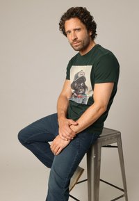Groene t-shirt met korte mouwen, met een grafische afbeelding van een hond in een pet, gelaagd over denim jeans. Model zit op een metalen kruk.