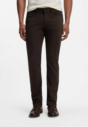 Mann trägt dunkelbraune gerade geschnittene Hose und dunkelbraune Schuhe, steht vor weißem Hintergrund.