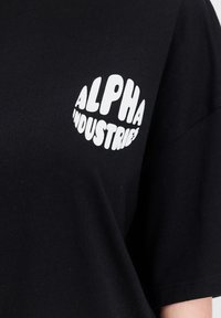 Černé bavlněné tričko s bílým kruhovým logem, které má nahoře nápis "ALPHA" a pod ním "INDUSTRIES" na levé části hrudi. Krátké rukávy, volný střih.