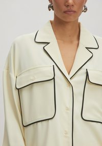 Crème blouse met zwarte piping, grote flapzakken en een notch-kraag. Gemaakt van een gladde stof met een relaxte fit en knoopsluiting.