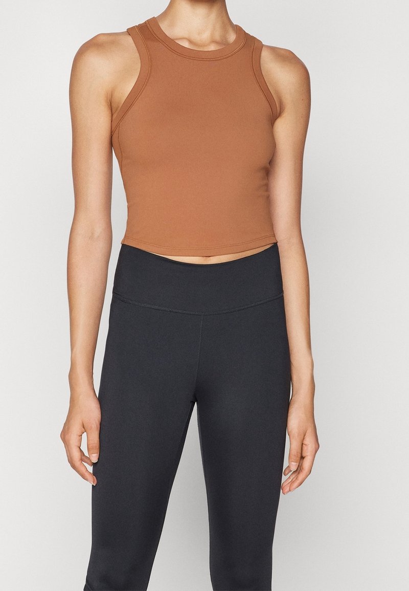 Femme portant un top court marron sans manches et des leggings noirs taille haute, debout devant un fond clair uni.