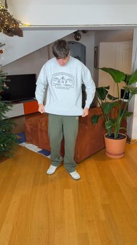 Γκρι φούτερ με λογότυπο μπροστά, φορεμένο με πράσινο παντελόνι και λευκά αθλητικά παπούτσια. Εσωτερικός χώρος με φυτό και ξύλινο πάτωμα.