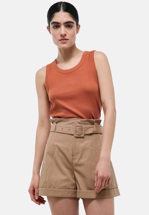 Jeune femme portant un haut sans manches couleur rouille et un short beige taille haute avec ceinture, debout devant un fond clair uni.