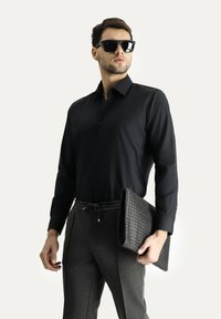 Camicia nera a bottone, pantaloni sportivi grigi con cordoncino, tiene una borsa nera texturizzata, indossa grandi occhiali da sole neri.